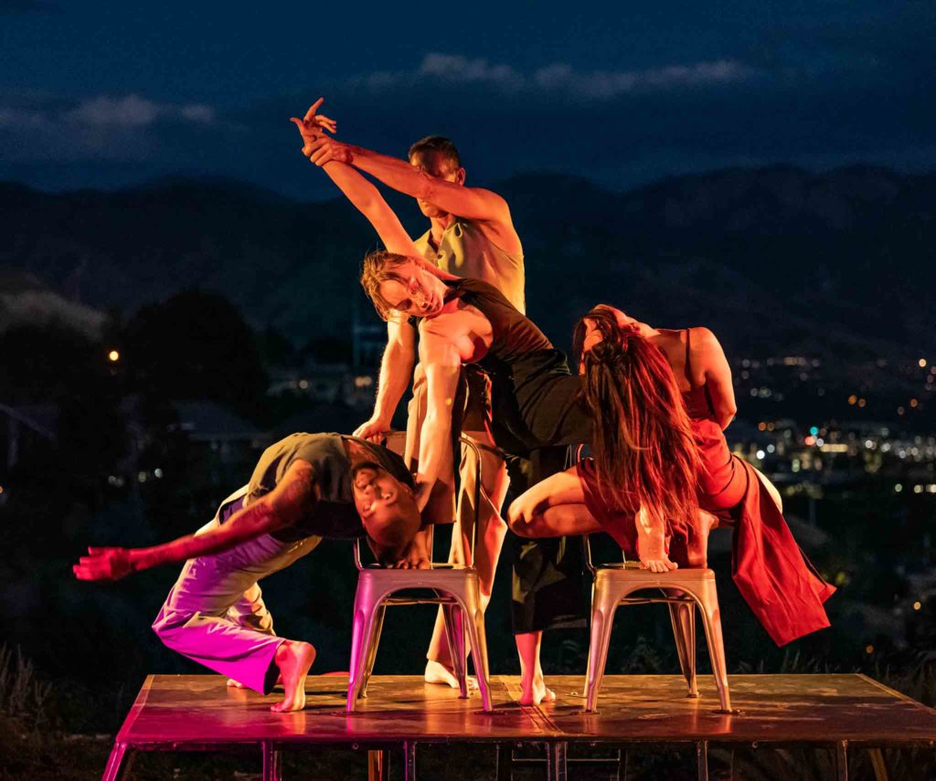 Four dancers on two chairs, each person in a different pose, against the backdrop of city lights at night